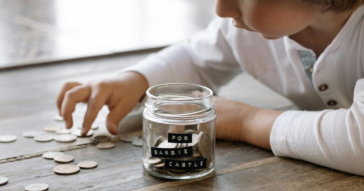 A young child counting coins on a kitchen table while a parent watches with a warm smile, soft natural light from a window.